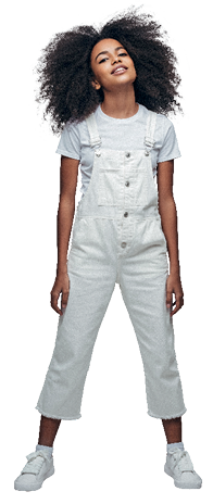 Imagem: Fotografia. Uma moça negra, de cabelos escuros Black Power, usando camiseta, jardineira e sapatos de cor branca. Ela olha para frente, com a cabeça um pouco inclinada para cima.  Fim da imagem.