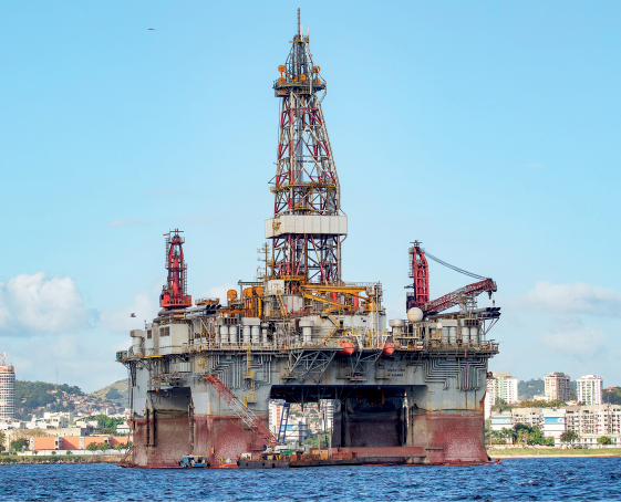 Imagem: Fotografia. Em alto mar, de água de cor azul, uma plataforma grande com base de cor cinza e rede de cor vermelha. Na parte superior, hastes de ferro na vertical. Em segundo plano, prédios, morros e cidades. Na parte superior, céu de cor azul-claro e nas pontas, nuvens de cor branca. Fim da imagem.