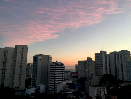 Imagem: Fotografia. Vista geral de cidade com muitos prédios, uns altos, outros médios, pertos uns dos outros. Na parte superior, céu de cor azul com nuvens de cor rosa-claro e partes em laranja.  Fim da imagem.