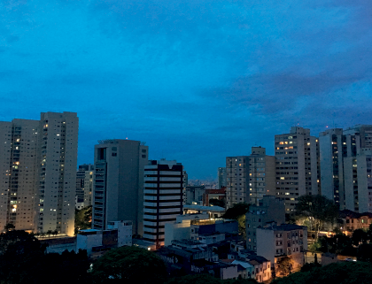 Imagem: Fotografia. A mesma cidade descrita anteriormente, com o céu de cor azul e com nuvens esparsas em cinza.  Fim da imagem.