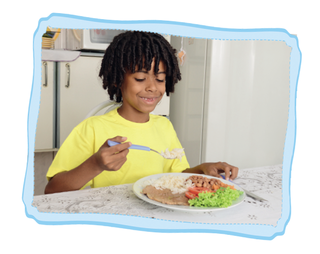 Imagem: Fotografia. Um garoto sentado de frente para uma mesa, de pele negra, de cabelos cacheados pretos, usando camiseta amarela. Ele segura um talher na mão esquerda e na mesa, um prato redondo branco com comida. Ele olha para ela sorrindo. Ao fundo, móveis da cozinha vistos parcialmente.  Fim da imagem.