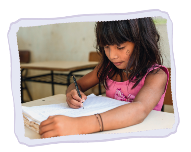 Imagem: Fotografia. Uma menina sentada de frente para carteira escolar, escrevendo com a mão esquerda sobre uma folha de cor branca. Ela tem pele morena, cabelos lisos com franja, escuros, usando uma blusa regata de cor rosa.  Fim da imagem.