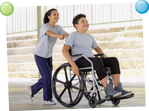 Imagem: Fotografia. Uma menina em pé de pele morena-claro, com cabelos pretos amarrados para trás, usando camiseta de cor cinza-claro, com calça azul e sapatos em branco com cadarço em preto. Ela empurra um menino cadeirante, de pele clara, cabelos escuros, com camiseta cinza-claro, com calça preta e sapatos cinzas. Ambos estão sorrindo.  Fim da imagem.