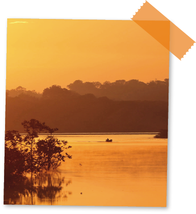 Imagem: Fotografia. O mesmo local descrito anteriormente, em tons de laranja no céu e reflexo no rio. Ao fundo, uma canoa com pessoa.  Fim da imagem.