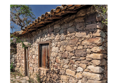 Imagem: Fotografia. À direita, casa com paredes feitas de pedras de cor marrom-claro, com janela pequena de madeira, porta do mesmo estilo e na parte superior, telhado marrom. Na parte superior, céu azul-claro e galhos finos de árvore com folhas verdes. Fim da imagem.