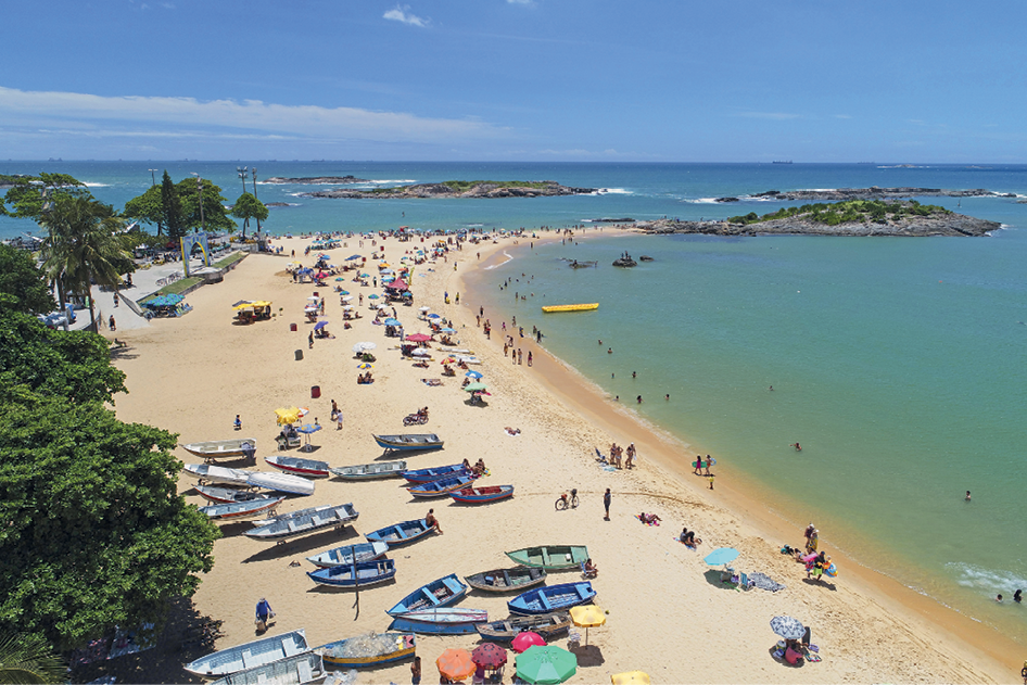 Imagem: Fotografia. Vista do alto de uma praia. À esquerda, vista parcial de árvores com folhas verdes. À frente delas, canoas e barcos pequenos de cor branca e outras em branco. Perto dos barcos, pessoas debaixo de guarda-sóis e outras pessoas de frente para praia. Mais à direita, mar com água de cor verde com pessoas se banhando na borda. Mais ao fundo, parte de terra com solo de cor cinza e vegetação verde. Na parte superior, céu de cor azul-claro com poucas nuvens.  Fim da imagem.