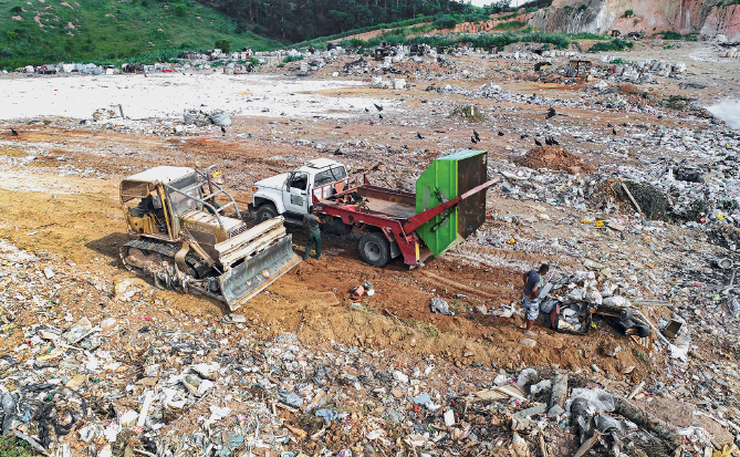 Imagem: Fotografia. Vista geral de local com solo de cor marrom, com centenas de dejetos, plásticos, garrafas e lixos. À esquerda, um trator de cor amarelo-claro, e pá à direita. Atrás, um caminhão de cor branca, fundo vermelho e caçamba de caminhão jogando lixo no solo. Em segundo plano, vista parcial de local com vegetação verde.  Fim da imagem.