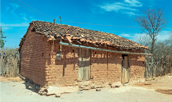 Imagem: Fotografia. Em um local com solo arenoso em bege, uma casa com tijolos de cor bege-claro, duas portas e telhado em cinza com o formato triangular. Atrás, local com vegetação seca e árvores com troncos finos, secas. Na parte superior, céu de cor azul-claro com nuvens brancas.  Fim da imagem.