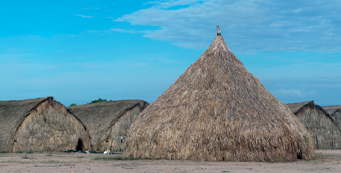 Imagem: Fotografia. Vista geral de local com solo de cor bege. Ao centro, uma grande cabana indígena com a forma arredondada e com a ponta triangular, coberta por palha grossa de cor bege e partes em cinza. Ao fundo, outras moradias menos, cobertas pelo mesmo tipo de palha, com o formato triangular. Na parte superior, céu de cor azul-claro e nuvens brancas na ponta da direita.  Fim da imagem.