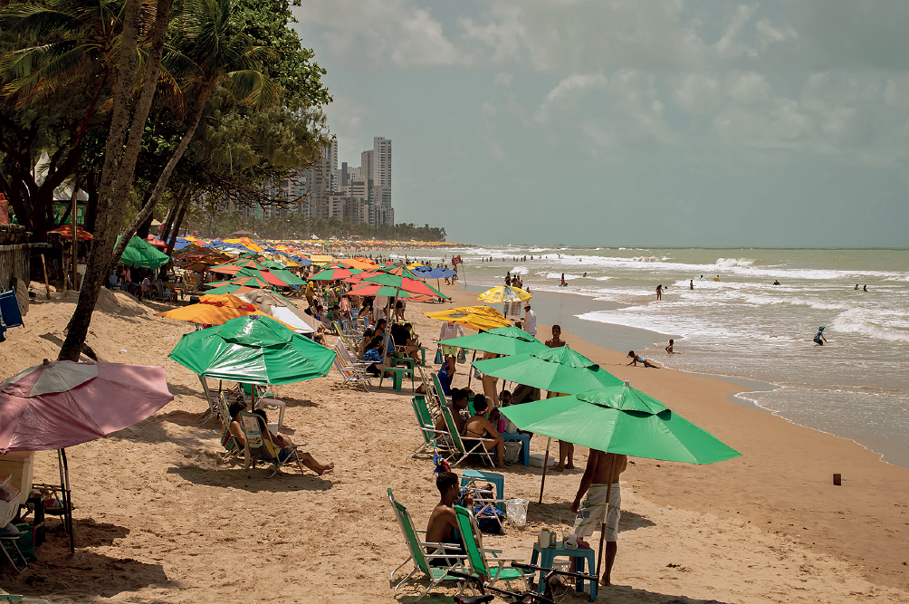 Imagem: Fotografia. À esquerda, praia com areia de cor bege-clara, onde há pessoas sentadas debaixo de guarda-sóis coloridos em verde, laranja, amarelo e vermelho. Na ponta da direita, algumas pessoas vistas parcialmente em pé, outras pessoas na beira da praia no mar. À esquerda, vista parcial de algumas árvores com folhas verdes. Ao fundo, prédios de cidade de paredes de cor cinza-claro. No céu, azul-claro com nuvens de cor branca.  Fim da imagem.