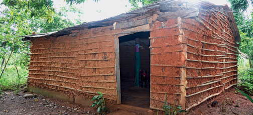 Imagem: Fotografia. Em um local com solo de cor marrom, ao centro, casa de tamanho médio, formato retangular com paredes de barro marrom, com partes em madeira. À frente, abertura de porta retangular na vertical. Ao fundo, local com muitas árvores com folhas verdes.  Fim da imagem.