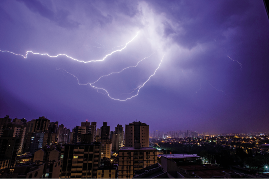 Imagem: Fotografia. Vista geral de cidade composta por prédios altos, com sombra sobre eles e na parte superior, céu noturno de cor azul-escuro com raio branco com ramificações saindo das nuvens.  Fim da imagem.