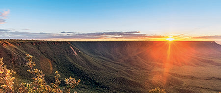 Imagem: Fotografia. Vista geral de local com morros altos com vegetação rasteira em verde. À direita, na parte inferior, debaixo de nuvens cinzas na horizontal, sol redondo em amarelo incandescente. Acima, céu de cor azul-claro. Fim da imagem.