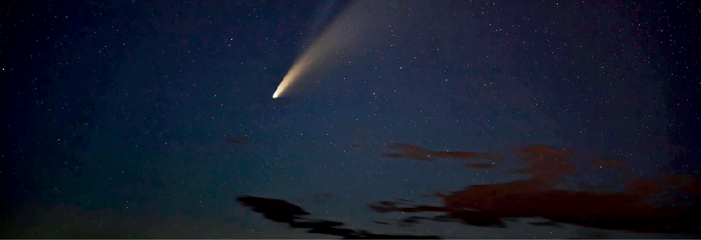 Imagem: Fotografia noturna. Céu de cor azul-escuro, com nuvens escuras na ponta inferior à direita, com pequenos pontos brancos coloridos. Na parte superior, cometa na vertical de cor branca com parte superior em bege.  Fim da imagem.