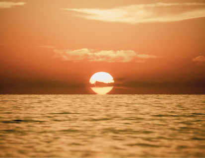 Imagem: Fotografia. Vista geral de local com mar, ao fundo, sol amarelo incandescente em meio a nuvens, no céu de cor alaranjada.  Fim da imagem.