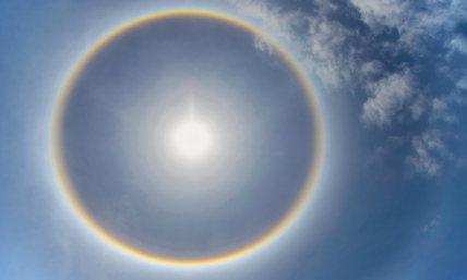 Imagem: Fotografia. Vista de baixo para cima, céu de cor azul-claro com nuvens brancas à direita. Ao centro, sol redondo com luz brancas incandescente com forma circular colorido em azul, amarelo e laranja.  Fim da imagem.