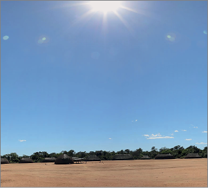 Imagem: Fotografia. Local com solo arenoso de cor bege, ao fundo, muitas casas com telhado triangular. Na parte superior, céu de cor azul-claro, com poucas nuvens brancas ao fundo e à frente, sol visto parcialmente com luz brancas incandescente.  Fim da imagem.