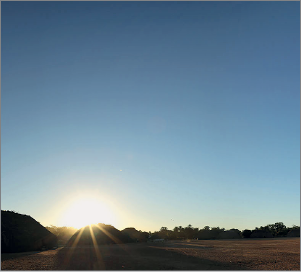 Imagem: Fotografia. O mesmo local descrito anteriormente, com sombra no solo e cidade, com céu em azul mais escuro, sol incandescente na parte inferior.  Fim da imagem.