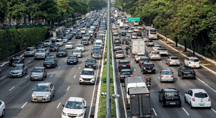 Imagem: Fotografia. Vista do alto de avenida em duas partes de cor cinza. À esquerda, dezenas de carros indo para frente. Ao centro, canteiro com grama verde e à direita, dezenas de carros em sentido contrários. Nos acostamentos, vegetação e árvores de folhas verdes.  Fim da imagem.