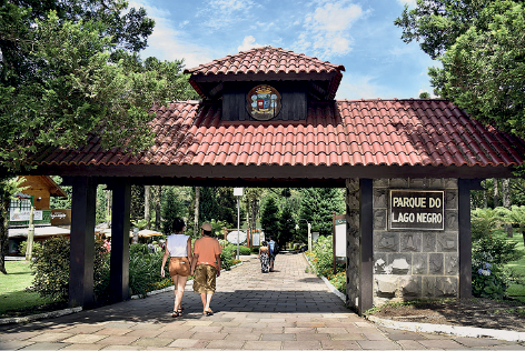 Imagem: Fotografia. Local aberto, com entrada de parede de cor cinza e na parte superior, telhado de cor marrom, com placa na parede à direita, em verde com texto branco : PARQUE DO LAGO NEGRO. À esquerda, entrada aberta, com duas pessoas passando vistas de costas, uma menina de pele clara, cabelos pretos, com blusa regata em branco, bermuda em marrom. À direita, outra pessoa, um homem de chapéu bege, camiseta de mangas curtas em laranja e bermuda em marrom. Mais ao fundo, local com vegetação verde e árvores. Na parte superior, céu azul-claro com nuvens brancas.  Fim da imagem.