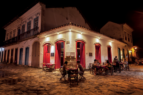 Imagem: Fotografia. No céu noturno em preto, com casarões de paredes brancas, com portas em vermelho. À esquerda e à direita, com dois andares, com janelas de cor azul. No teto, luz branca. De frente para as portas, mesas pequenas com cadeiras e pessoas comendo.  Fim da imagem.