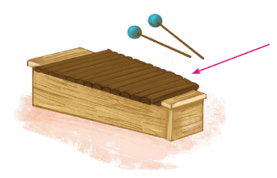 Imagem: Um xilofone, instrumento de cor bege com tubos finos na horizontal em marrom-escuro um ao lado do outro. Na parte superior, duas baquetas com ponta superior redonda em azul. Uma seta indica os tubos. Fim da imagem.