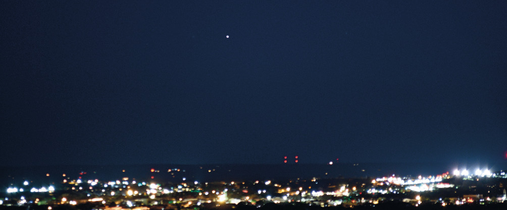 Imagem: Fotografia. Na parte inferior, cidade iluminada com luzes acesas em amarelo e tons de laranja. Na parte superior, céu noturno em azul-escuro e ao centro, uma estrela brilhante de cor branca. Fim da imagem.