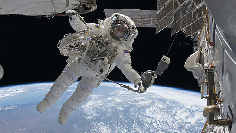 Imagem: Fotografia. Um homem com roupa de astronauta, cobrindo braços e pernas e sobre a cabeça, capacete. Ele está sobrevoando no espaço, perto de uma nave espacial. Na parte inferior, vista parcial de esfera terrestre em azul, com nuvens brancas.  Fim da imagem.
