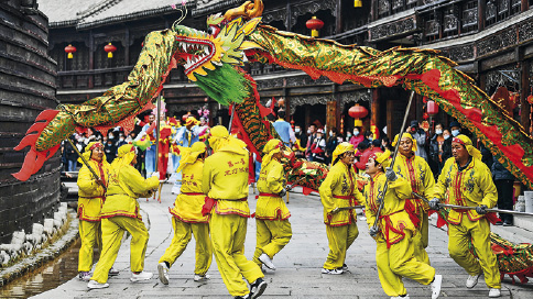 Imagem: Fotografia. Em uma ala de cor cinza-claro, com dezenas de pessoas com roupa de mangas compridas e calça em amarelo, com detalhes em vermelho. Eles estão com hastes finas na vertical, carregando no alto um dragão. Este tem o corpo grande, com o corpo na horizontal, dourado, com partes em vermelho, com à cabeça para à esquerda, boca aberta, dentes brancos e contorno da boca em laranja. Mais ao fundo, outras dezenas de pessoas.  Fim da imagem.