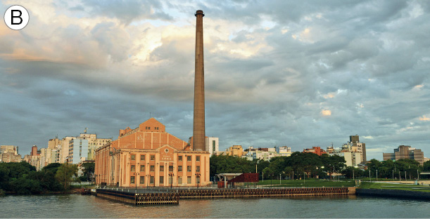 Imagem: Fotografia B. Vista geral de local com rio à frente, com porto de cor marrom-escuro e em segundo plano, edifício grande de cor marrom-claro, com muitas janelas. À direita, na vertical estrutura gigante de cor marrom. Em segundo plano, vegetação com folhas verdes e mais ao fundo, cidade composta com prédios de tamanhos diversos. Na parte superior, céu azul-claro com inúmeras nuvens brancas.  Fim da imagem.