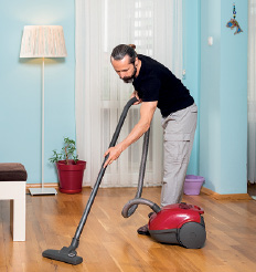 Imagem: Fotografia. Um homem passando aspirador no chão. Ele tem pele clara, cabelos e barba em preto, camiseta da mesma cor, calça cinza e aspirador em vermelho. Ao fundo, parede em azul com abajur grande, janela e cortina branca sobre ela.  Fim da imagem.