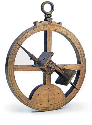 Imagem: Fotografia. Um astrolábio, objeto de madeira com formato redondo, com círculo pequeno na ponta superior, com haste na vertical e na horizontal. Sobre essa forma, ferro na vertical, com ponta fina na parte superior e parte inferior.  Fim da imagem.