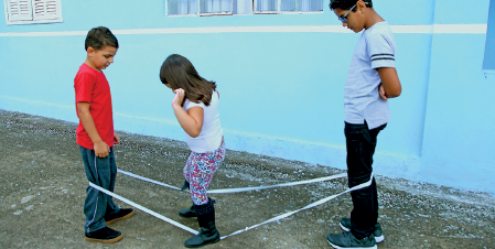 Imagem: Fotografia. Três crianças brincando em local com solo cinza e paredes em azul-claro. À esquerda, menino de pele morena, cabelos castanhos com camiseta vermelha, calça azul e sapatos pretos, com elástico branco passando pelas pernas dele. Ao centro, menina para à esquerda, pisando em elástico. Ela tem pele clara, cabelos castanhos nos ombros, com blusa de mangas curtas, calça estampada em rosa e azul e botas pretas. À direita, outro menino, maior, de pele morena, cabelos pretos, com par de óculos no rosto, com camiseta de mangas curtas em cinza, calça preta e sapatos em verde e branco. A outra ponta do elástico passa sobre as pernas dele.  Fim da imagem.