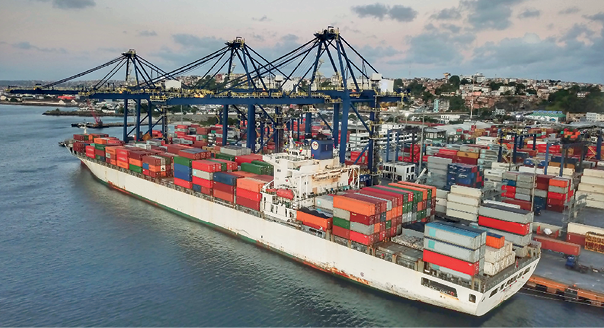 Imagem: Fotografia. Vista do alto para baixo, em rio grande com água escura, um navio grande de carga de cor branca, com contêineres na horizontal, aglomerados coloridos. Ao fundo, outros navios com contêineres e hastes de ferros em preto e parte em marrom na horizontal. Em segundo plano, cidade com casas e morros com vegetação verde. Na parte superior, azul, com nuvens.  Fim da imagem.