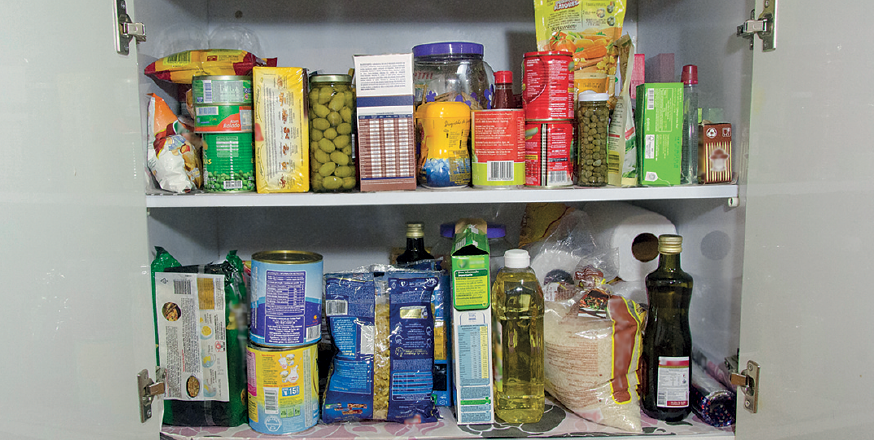 Imagem: Fotografia. Dentro de um armário, alimentos em embalagens plásticas, caixas de papelão, latinhas redondas e pequenas, pote de vidro com conserva de azeitonas verdes, garrafa plástica com óleo e uma garrafa de vidro com azeite. Mais ao fundo, na parte de baixo, rolos de guardanapo branco.  Fim da imagem.