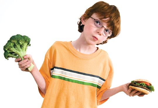 Imagem: Fotografia. Um menino visto da cintura para cima, com cabelos castanhos, com óculos de grau no rosto, com camiseta laranja, com listra na horizontal em branco e verde. Ele segura na mão esquerda, um pé de brócolis de cor verde e na mão direita, sanduíche com hamburguer, salada com tomate, olhando para o alto, à esquerda.  Fim da imagem.