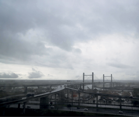 Imagem: Fotografia. Vista geral de local com céu de cor cinza, nublado. Na parte inferior, cidade com ruas, automóveis e ponte grande com duas hastes na vertical, estaiada.  Fim da imagem.