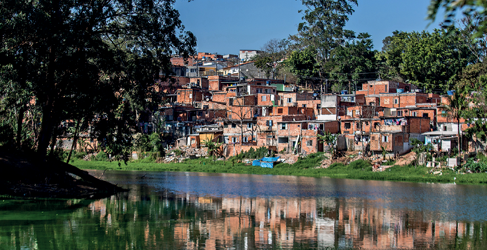 Imagem: Fotografia. Vista geral de local com rio de água escura, com bordas com vegetação, à esquerda, árvores com folhas verdes e à direita, ao fundo, inúmeras casas de paredes de bloco de cor laranja, amontoadas. Elas estão inacabadas, com formas diversas, sobrepostas entre si. Na parte superior, céu azul-claro.  Fim da imagem.