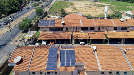 Imagem: Fotografia. Vista do alto de casas com telhados de cor marrom e na ponta da esquerda, placas solares de cor cinza-escuro. As placas estão dispostas na horizontal e na parte vertical. À esquerda, rodovia com árvores de folhas verdes. Ao fundo, solo de cor bege com vegetação verde.  Fim da imagem.