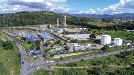 Imagem: Fotografia. Vista geral de local com formas arredondados grandes, branca, cinza e ao fundo, galpões e grande estruturas na vertical de cor cinza. À esquerda, outros galpões de cor azul. Ao redor do local, vegetação rasteira de cor verde. Em segundo plano, árvores altas de folhas verdes, morros e na parte superior, céu de cor azul-claro, com nuvens brancas.  Fim da imagem.