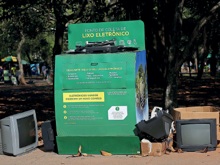 Imagem: Fotografia. Em local aberto com solo bege, com árvores com tronco marrom e folhas verdes na parte superior. À Frente, um grande contêiner verde na vertical com textos e na parte superior, perto da abertura, título:  Ponto de coleta de lixo eletrônico. Na abertura, aparelhos eletrônicos de cor preta. Sobre o solo, à esquerda, uma televisão de cor cinza, à direita, uma televisão em cinza-escuro, outros aparelhos e caixotes de cor bege.  Fim da imagem.