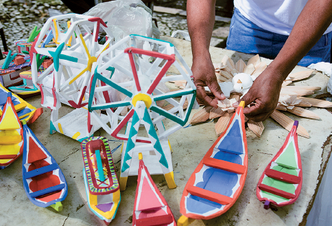 Imagem: Fotografia. Vista de cima para baixo, base de cor bege-claro, com objetos expostos:  pequenos barcos coloridos:  contornos em vermelho, laranja e azul e parte interna em verde, azul e vermelho. Ao fundo, à esquerda, duas rodas gigantes, de formato miniatura com detalhes em vermelho, verde com partes em amarelo. Ao fundo, à direita, vista parcial de braços de homem de pele morena, com camiseta branca e calça azul.  Fim da imagem.