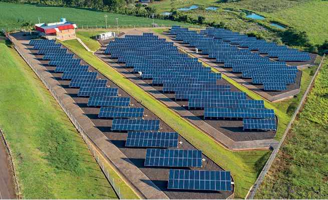 Imagem: Fotografia. Vista geral do alto, solo de cor cinza, com quatro fileiras na horizontal, com dezenas de placas solares de cor cinza-escuro, uma atrás da outra. À esquerda, estabelecimento de paredes de cor marrom-claro, com parte superior com detalhes em vermelho. Ao redor, vegetação rasteira de cor verde.  Fim da imagem.