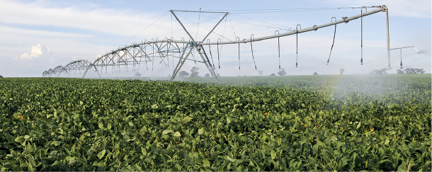 Imagem: Fotografia. Vista geral de local com vegetação com folhas verdes e ao fundo, uma estrutura de ferro na horizontal, com pontas para baixo e ao centro, estrutura com formato de dois triângulos, um normal e outro invertido em cima. Nessa estrutura, gotículas de água. Na parte superior, céu azul-claro com nuvens brancas.  Fim da imagem.