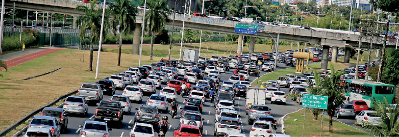 Imagem: Fotografia. Vista do alto de local com rodovia cinza com cinco faixas na vertical, com muitos carros, vistos pela parte traseira. Na parte superior, ponte cinza com outros carros passando. À esquerda e à direita, local com vegetação seca, com árvores de folhas verdes.  Fim da imagem.