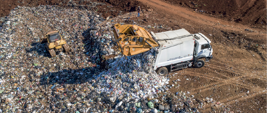 Imagem: Fotografia. Vista do alto para local com terreno aberto, à direita, solo de cor marrom, com um caminhão de cor branca, abrindo o fundo do caminhão soltando dejetos sobre o solo. Mais à esquerda, solo com centenas de dejetos e um trator menor de cor amarela, com rodas cinzas.  Fim da imagem.