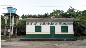 Imagem: Fotografia. Um estabelecimento de paredes de cor branca, com janelas e porta em verde, com o título: Quilombola de Mangabeira. No alto, telhado de cor marrom. À esquerda, sobre estrutura cinza na vertical, caixa em azul-claro. Em segundo plano, local com árvores de folhas em verde e céu em azul-claro.  Fim da imagem.