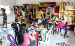 Imagem: Fotografia. Vista geral de local com paredes e pisos brancos, com uma estante de cor amarela com livros dentro, ao fundo. Ao centro, duas mesas de plástico de cor roxa, com cadeiras e crianças sentadas. À esquerda, uma mulher em pé, morena, cabelos escuros presos para trás, roupa colorida em amarelo e azul. De frente para a estante, uma criança vista de costa, com camiseta branca. À direita, uma mulher vista de costas de blusa amarela de mangas curtas.  Fim da imagem.