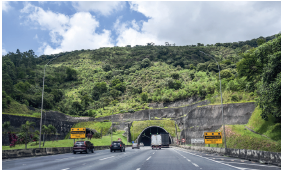 Imagem: Fotografia. Vista geral de local com rodovia com três faixas, por onde passam dois carros de cor preta e um caminhão, vistos de costas. Mais ao fundo, uma grande rocha, com túnel construído embaixo. Sobre a rocha e nos entornos à esquerda e à direita, vegetação rasteira e árvores com folhas verdes. No alto, céu azul com nuvens.  Fim da imagem.