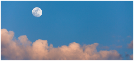 Imagem: Fotografia. Céu de cor azul-claro com nuvens de cor bege-claro na ponta inferior à esquerda e mais acima, lua redonda de cor branca.  Fim da imagem.